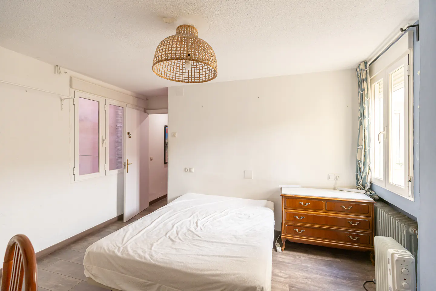 A bright bedroom with white walls, a double bed, a wooden dresser, and a woven pendant light. A window with patterned curtains is on the right.