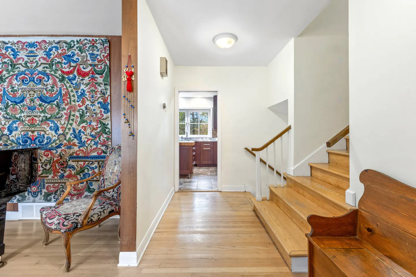 Foyer with light wood floors, stairs, and a bench. A colorful tapestry hangs near a chair and a glimpse of the kitchen is visible.