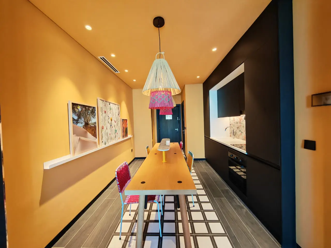 A modern apartment interior with yellow walls, a long wooden table with colorful chairs, and a black kitchen area. A unique, tiered pendant light hangs above the table.