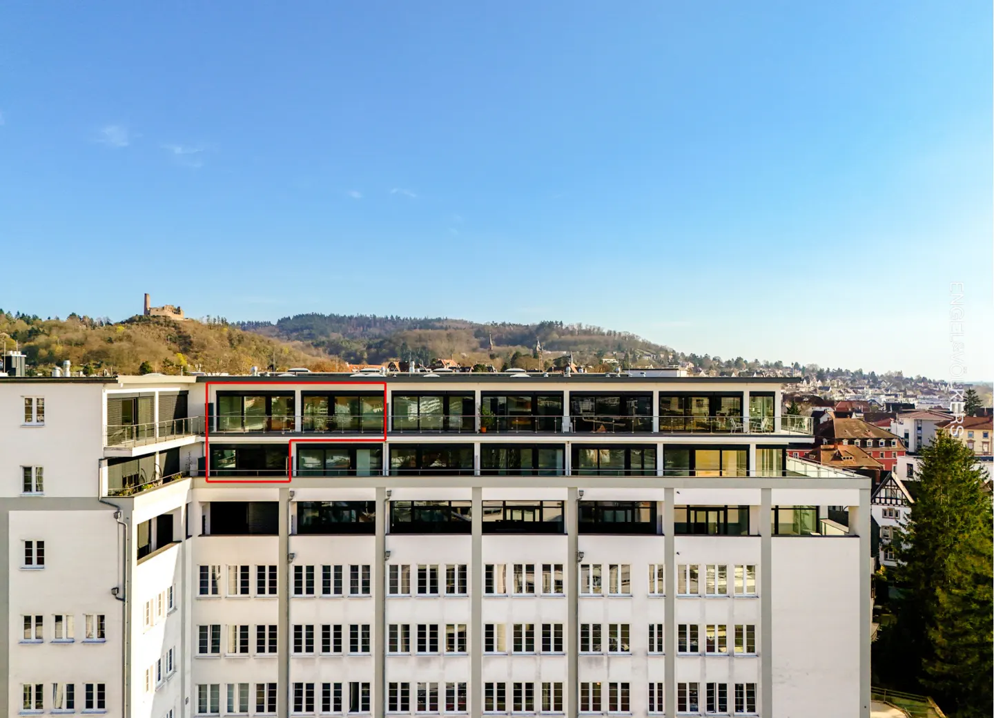 Exterior of a modern white apartment building with balconies and large windows, set against a blue sky and distant hills.
