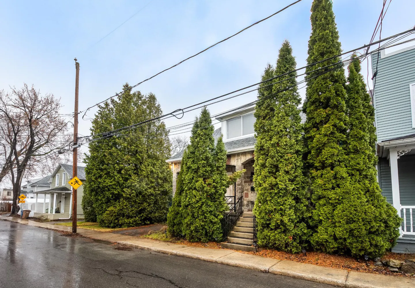 Street view of a two-story house with tall green trees in front. The sky is overcast, and the road is wet.