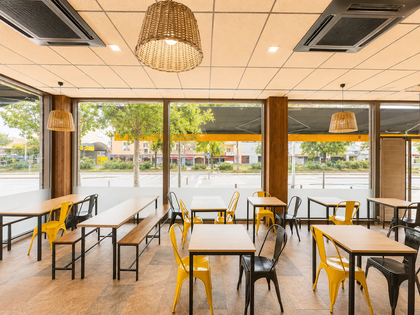 Modern cafe interior with wooden tables, yellow and black metal chairs, wicker pendant lights, and large street-view windows.