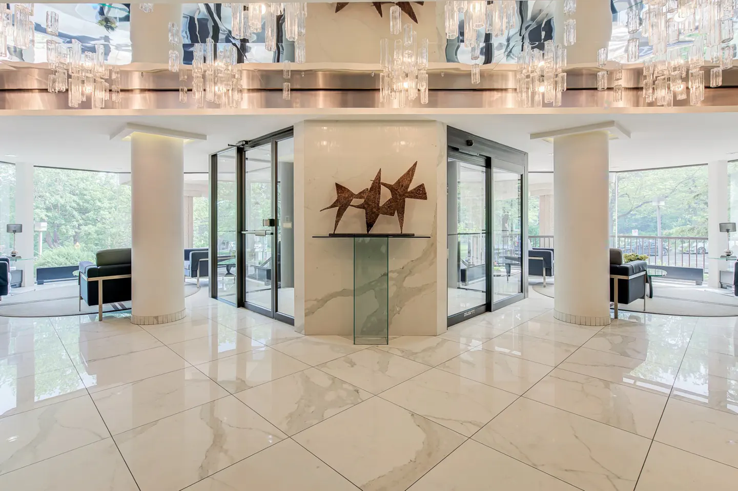 Lobby with marble floors, columns, and glass doors. A metal bird sculpture sits on a glass stand. Chandeliers hang from the ceiling.