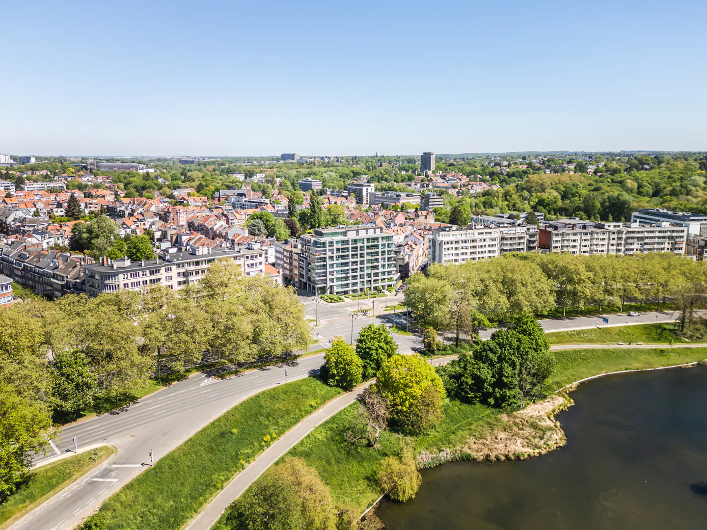 Aerial view of a city with red-roofed houses, modern apartments, green trees, a road, and a lake under a clear blue sky.