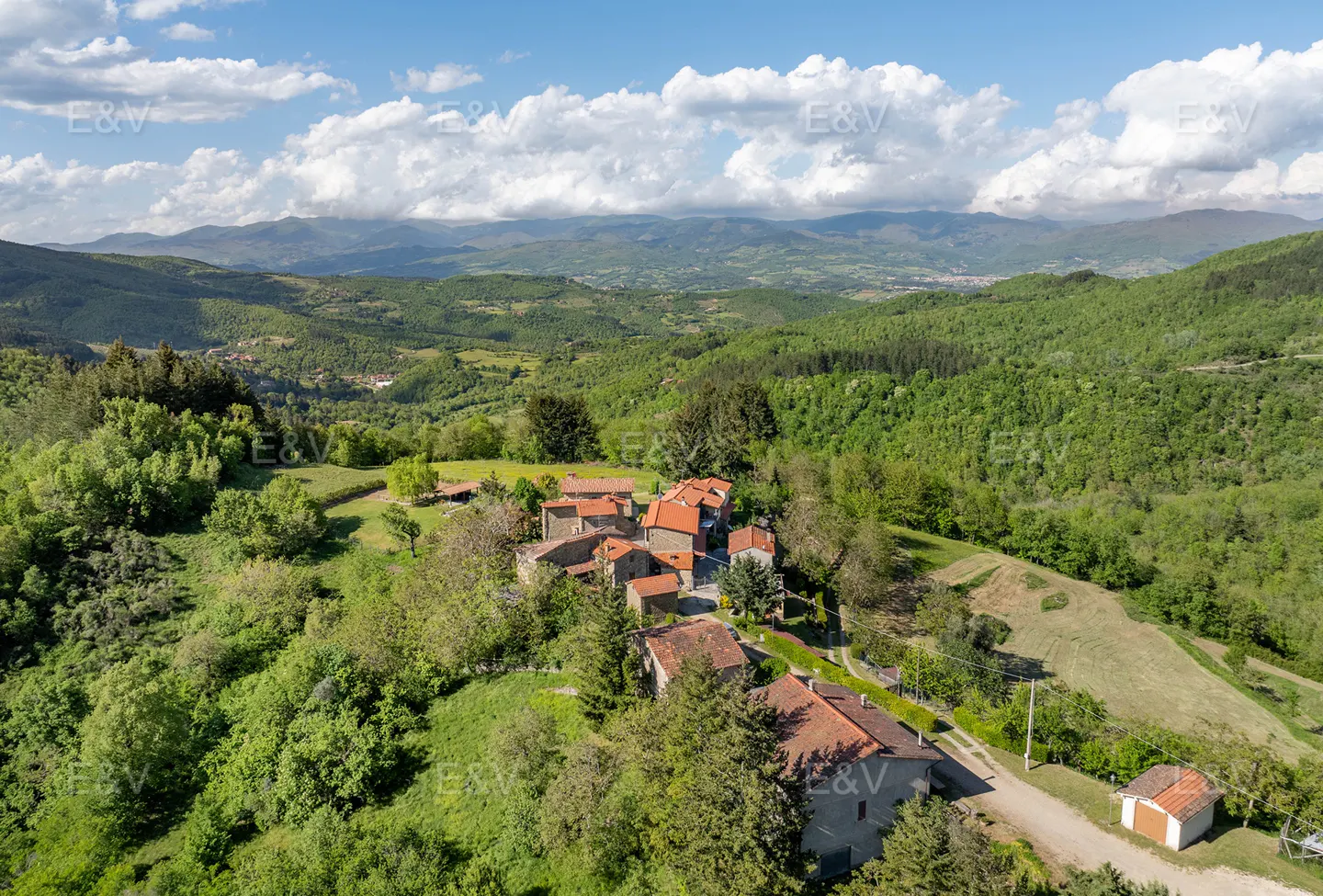 Aerial view of a Tuscan village with red-tiled roofs, surrounded by green hills and a blue sky with white clouds.