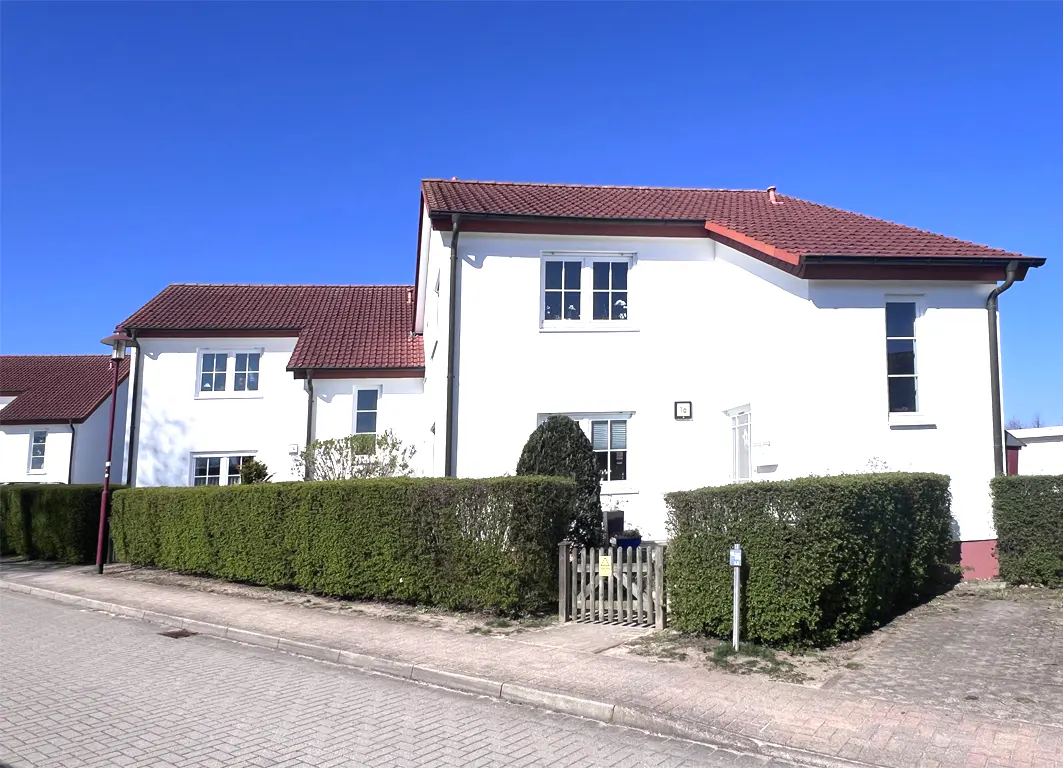 A white house with a red roof is partially obscured by a green hedge under a clear blue sky.