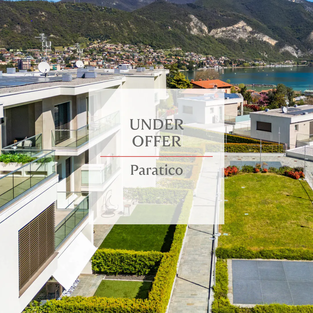 Aerial view of modern white apartments with glass balconies, green lawns, and a lake and mountains in the background. "Under Offer Paratico" text overlay.