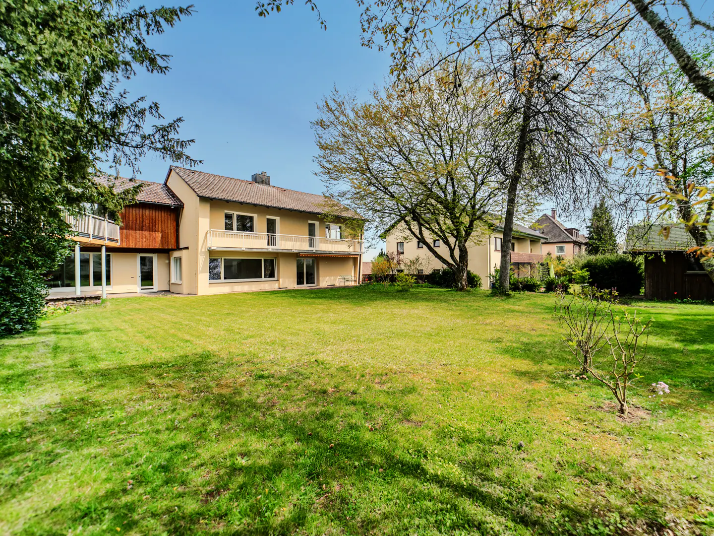 A tan two-story house with a large green lawn and trees under a blue sky.