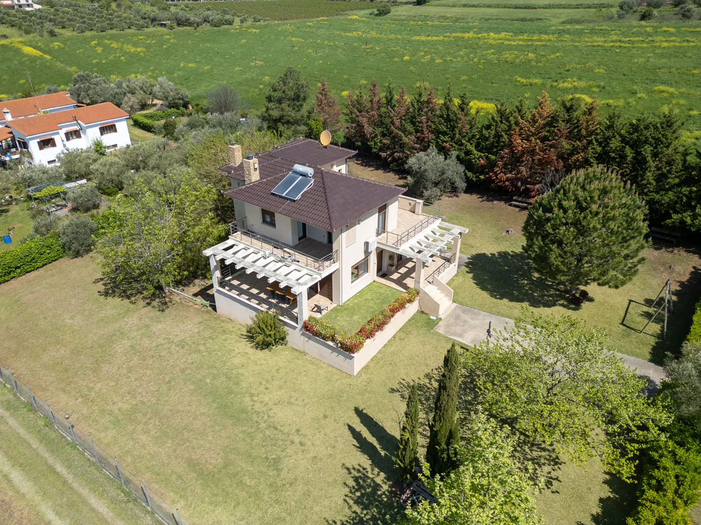 Aerial view of a two-story beige house with a brown roof, solar panel, and pergola in a green lawn with trees.