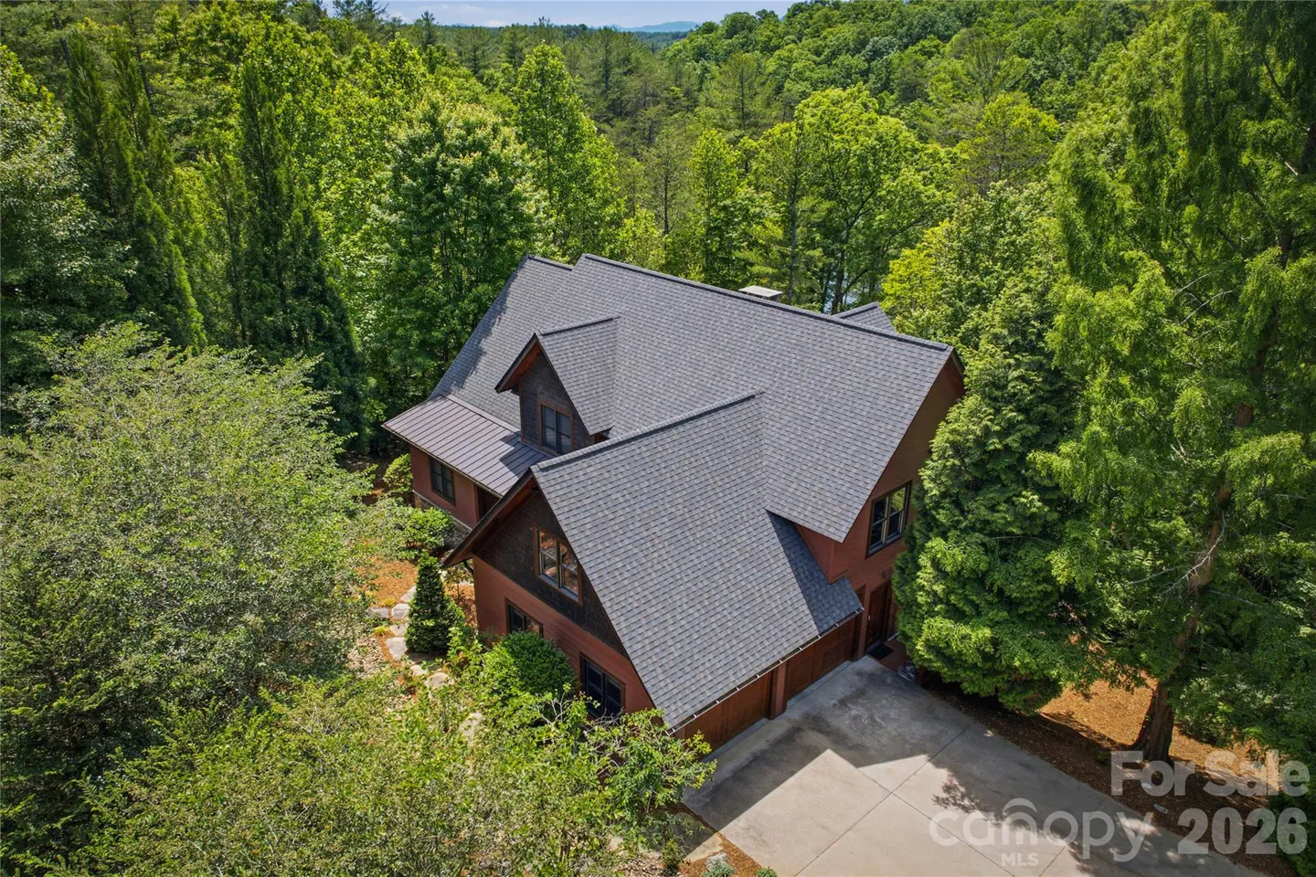 Aerial view of a brown house with a gray roof surrounded by green trees. A concrete driveway leads to the garage.