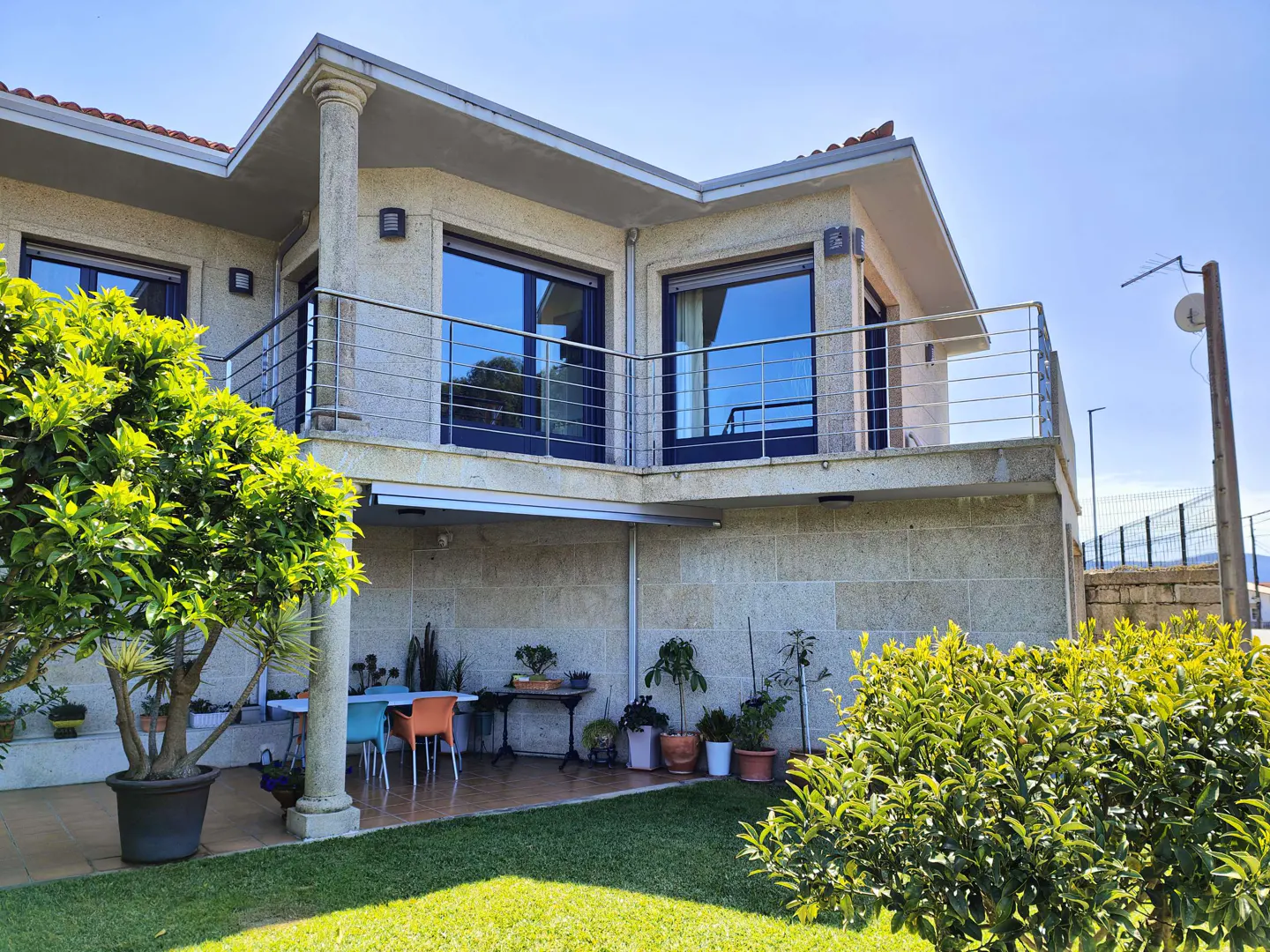 Two-story stone house with a balcony, lawn, and patio furniture on a sunny day.