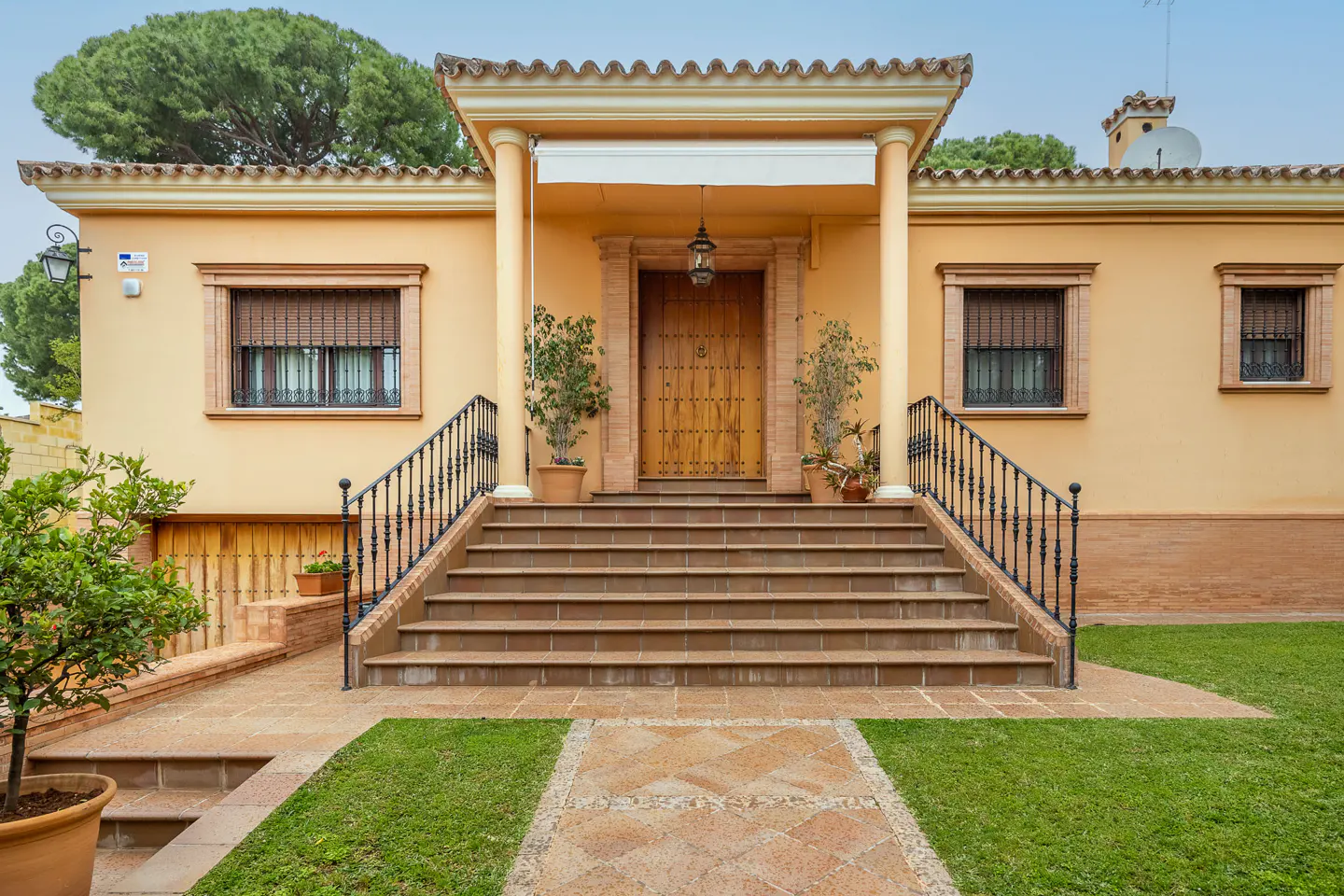 Tan house with a large wooden door, columns, and a long staircase with black railings. Green grass and trees surround the house.