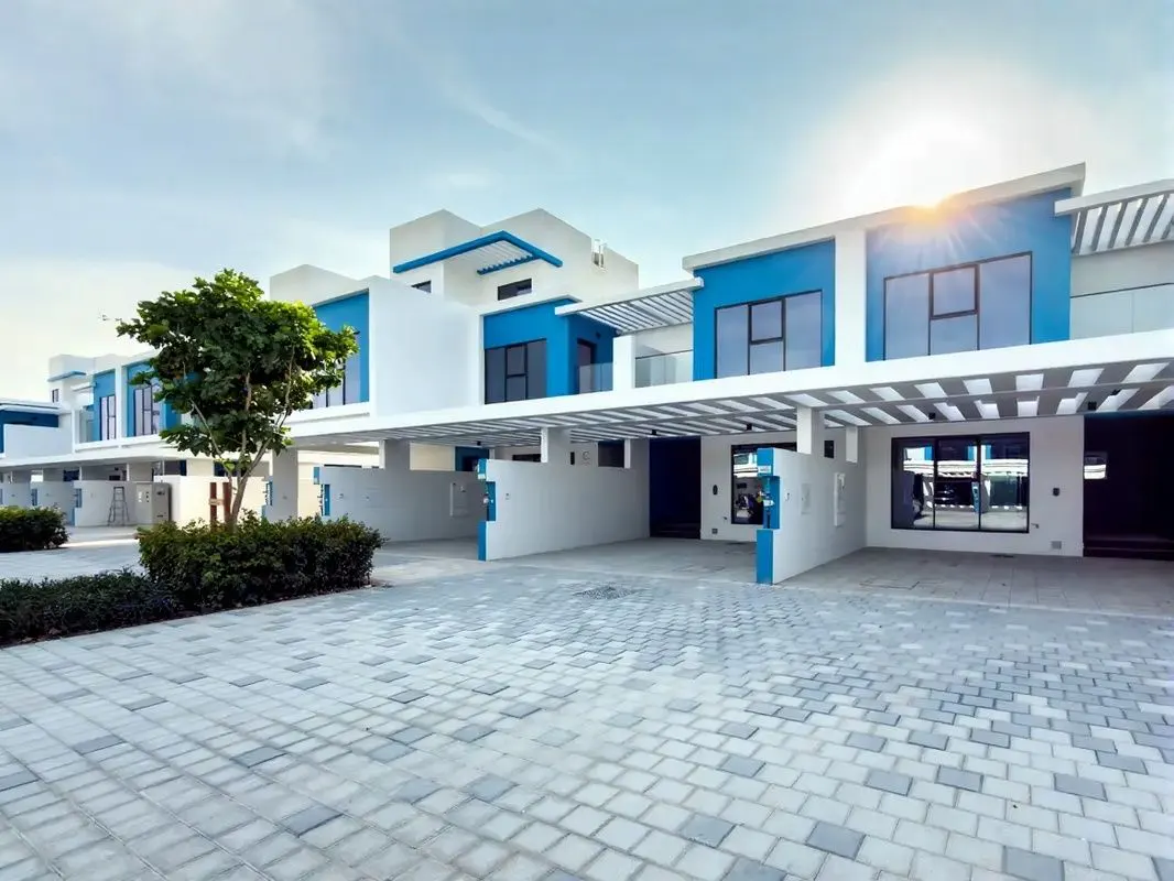 Row of modern, two-story townhouses with white and blue exteriors, carports, and a paved driveway under a sunny sky.