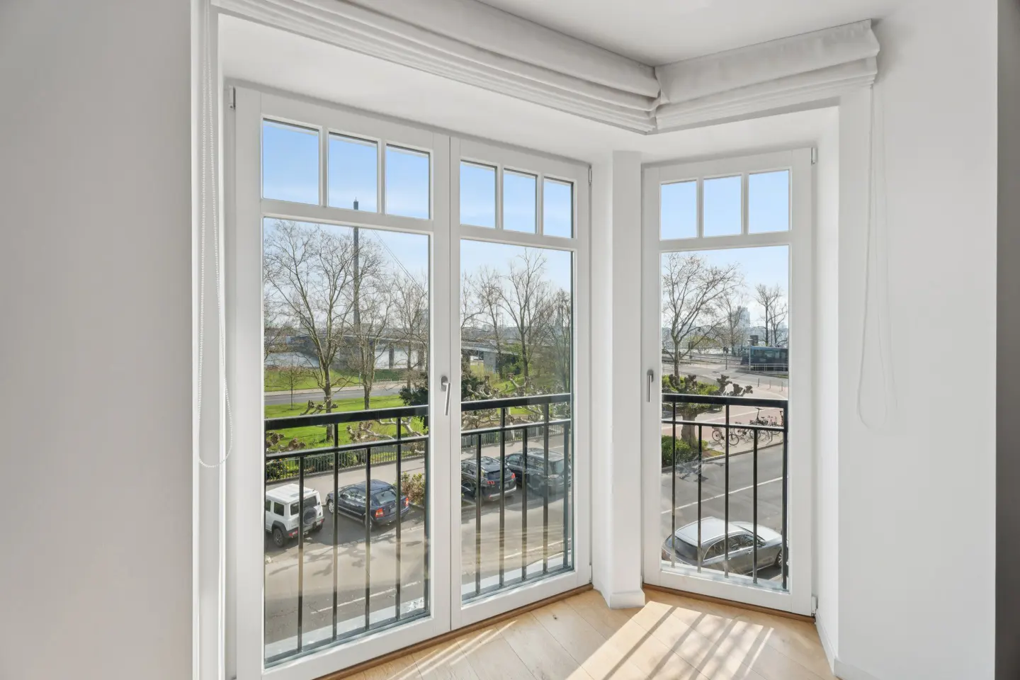 Bright room with white trim windows and a balcony overlooking a park with trees, cars, and a bridge in the distance.