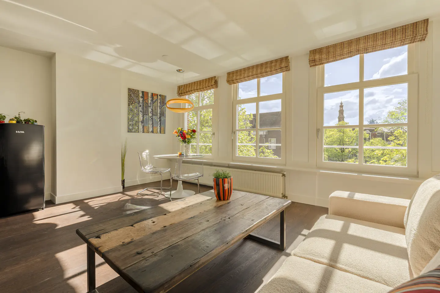 Bright living room with wood floors, white walls, and large windows with woven shades. A rustic wood coffee table sits in front of a beige sofa. A black fridge is on the left.