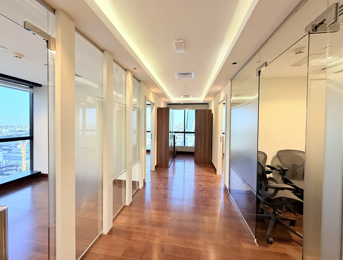 Bright office hallway with wood floors, glass-walled offices, and a city view through large windows at the end.