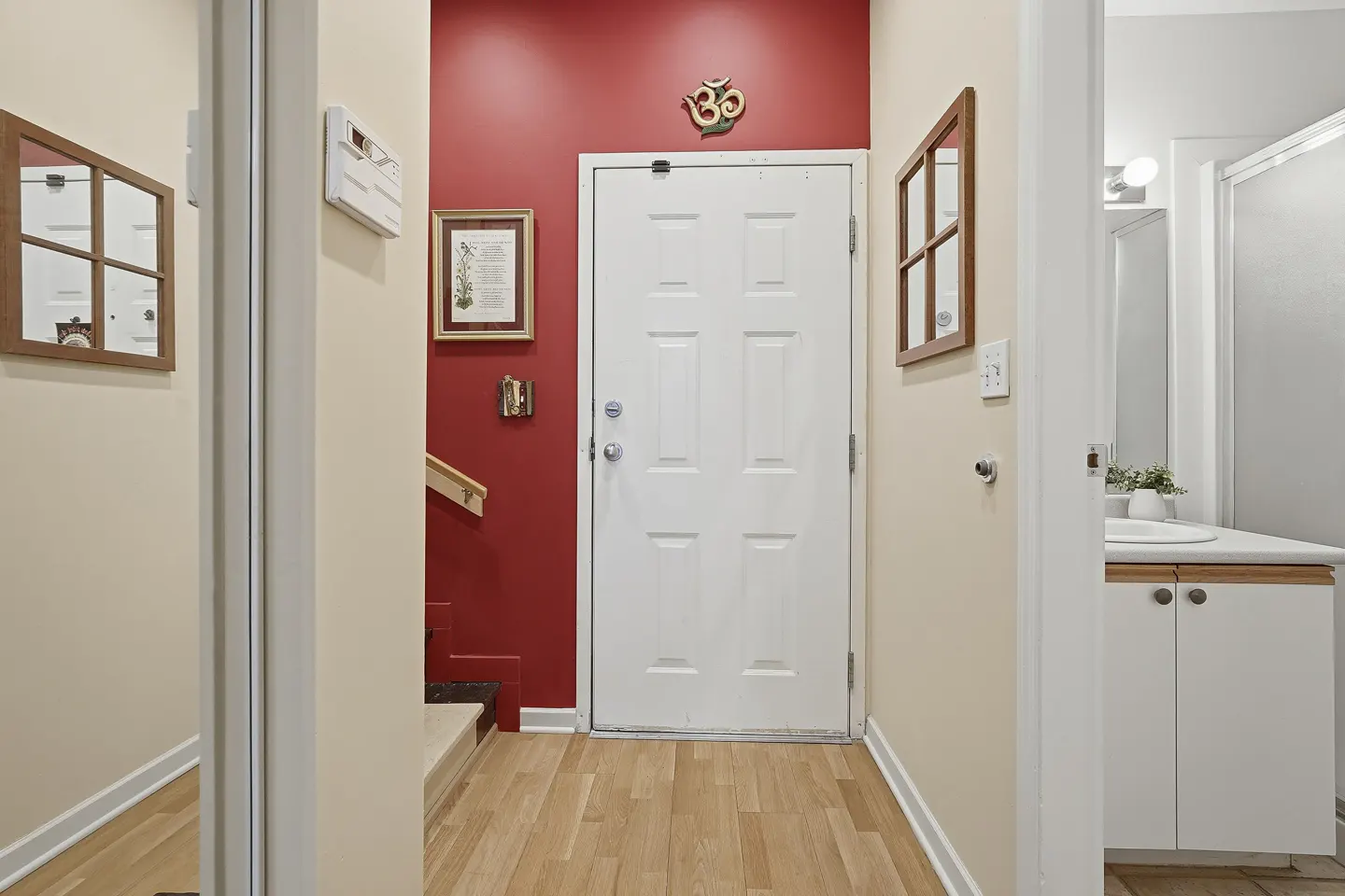 Entryway with a white door, red accent wall, light wood floors, and a view into a bathroom with a white vanity.