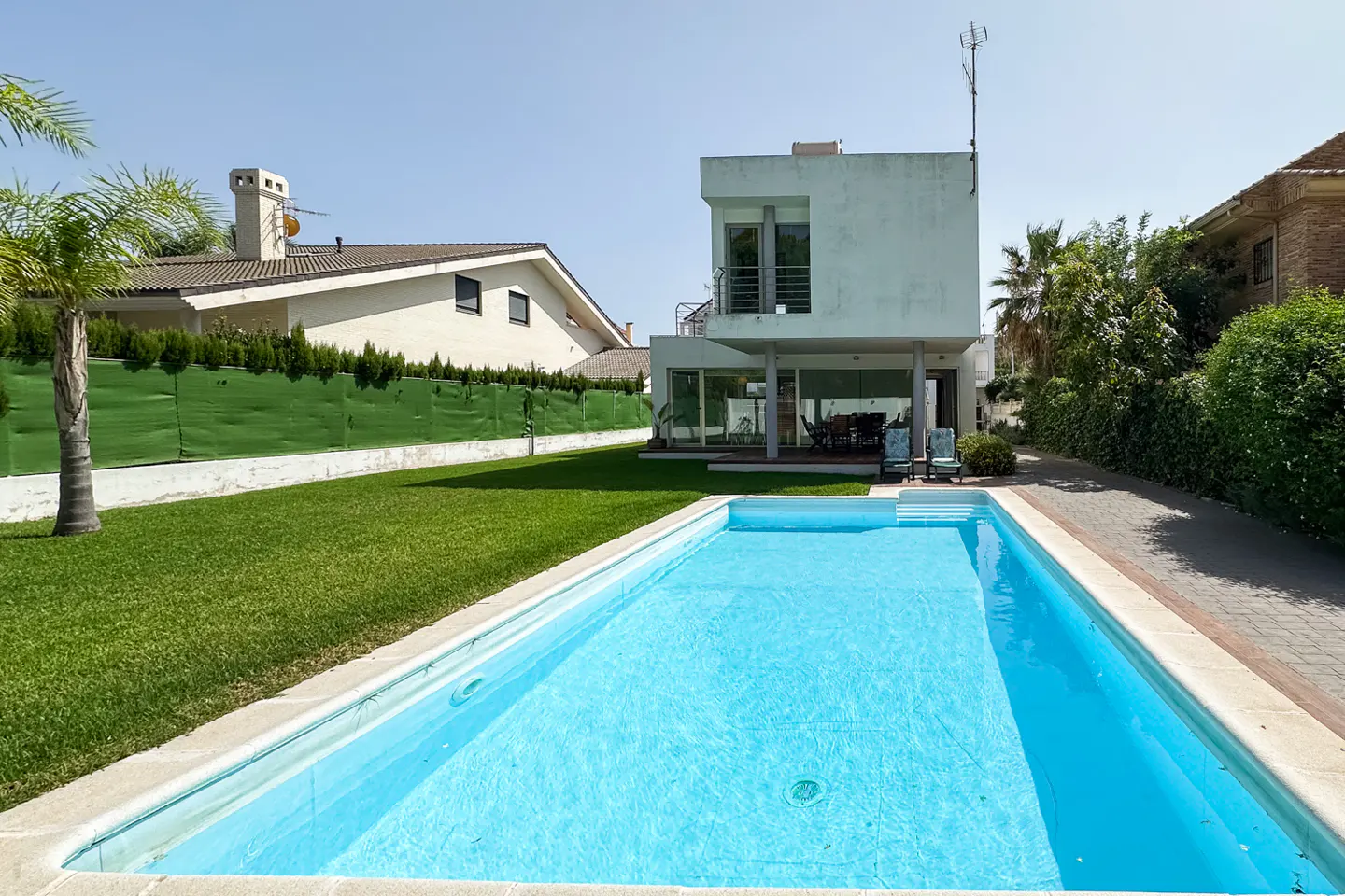 A modern two-story house with a rectangular blue swimming pool, a green lawn, and a palm tree under a clear blue sky.