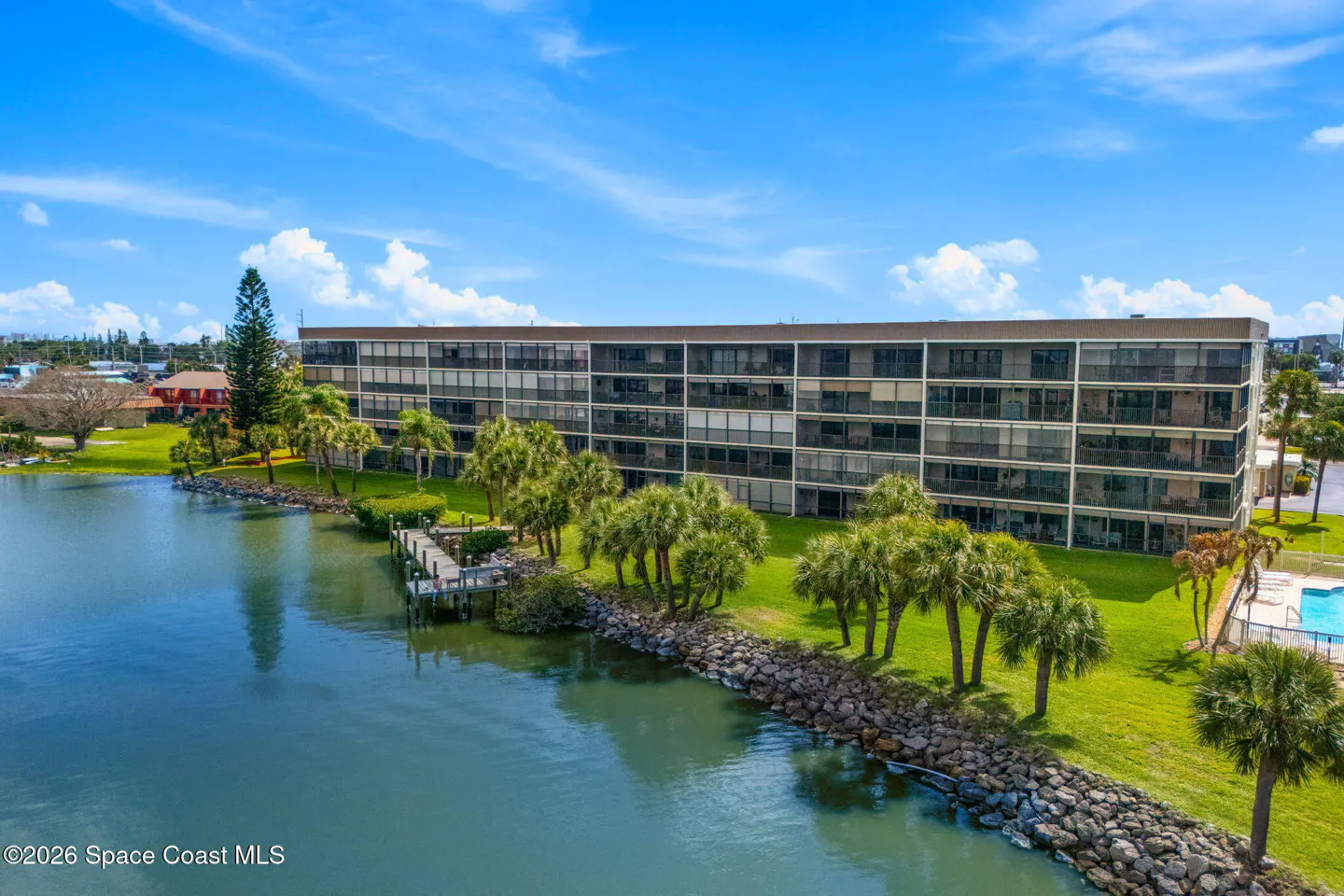 Aerial view of a waterfront condo building with a dock, palm trees, and a blue sky.