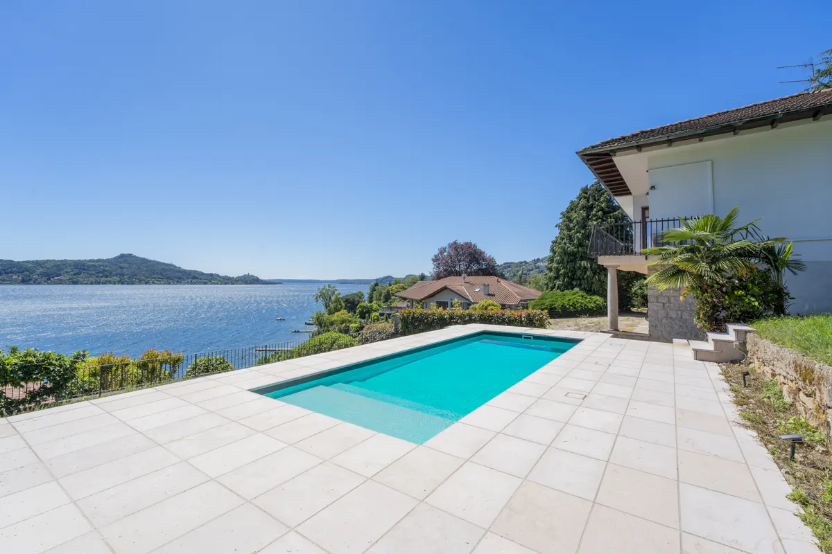A rectangular turquoise pool sits on a white tiled patio overlooking a blue lake and sky. A white house is on the right.