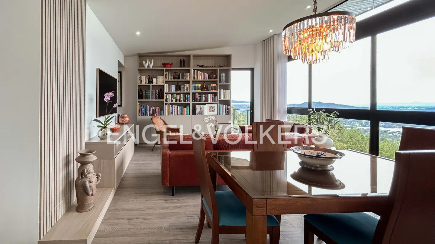 A bright living room with a dining table, a red sofa, and a bookshelf. A chandelier hangs above the table, and a large window shows a view of the mountains.