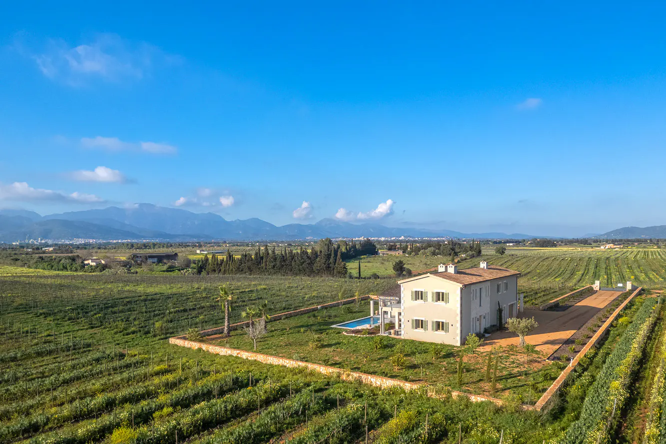 Aerial view of a two-story beige house with a pool, surrounded by vineyards under a blue sky. Mountains are visible in the distance.