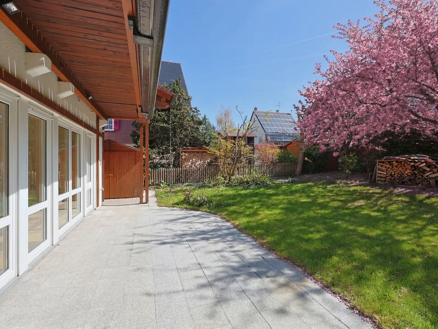A backyard with a stone patio, green grass, and a blooming pink tree. A wood fence and houses are in the background.