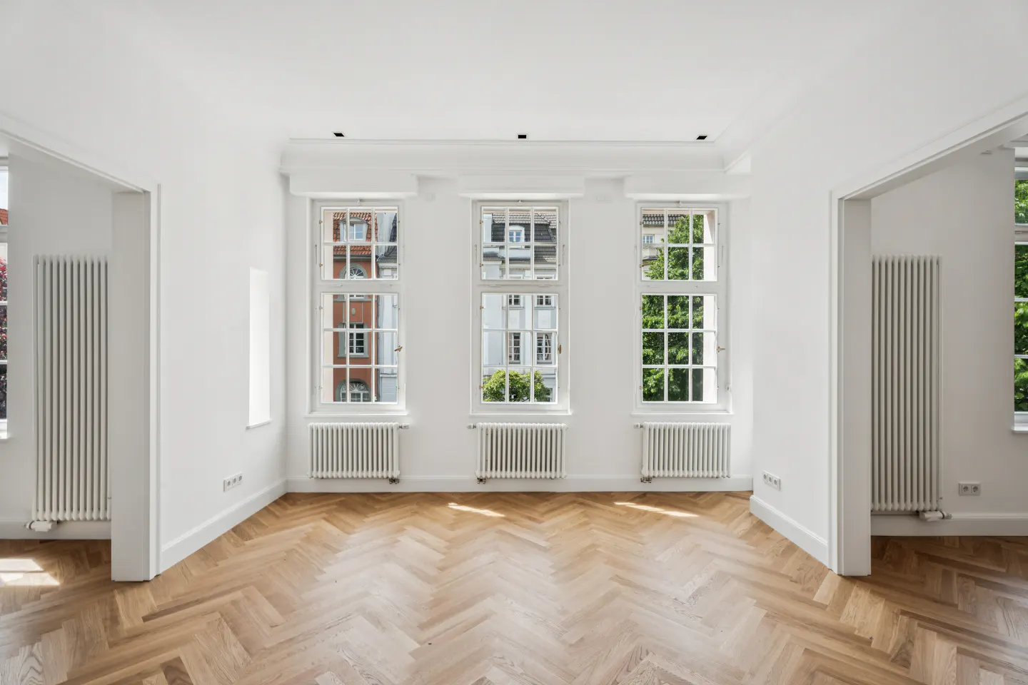 Bright, empty room with white walls, herringbone wood floors, three large windows, and white radiators.