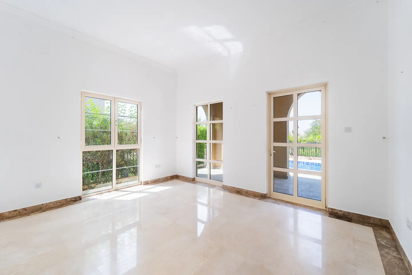 Bright, empty room with white walls, marble floors, and three glass doors/windows. Outside, greenery and a pool are visible.