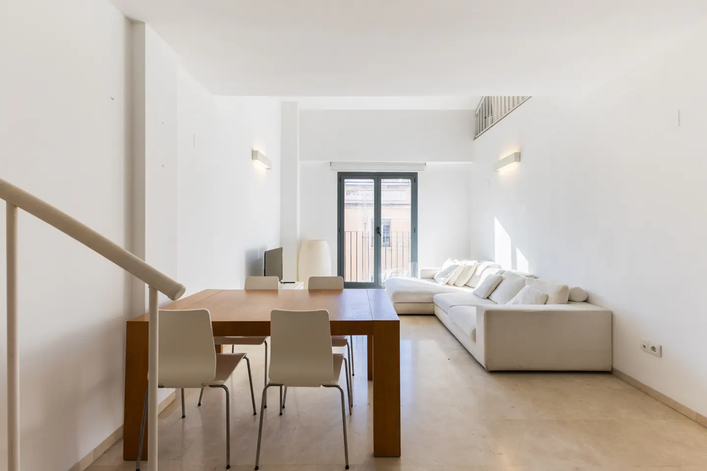 Bright, modern living room with a wooden table, white chairs, and a large white sectional sofa. A balcony door lets in natural light.