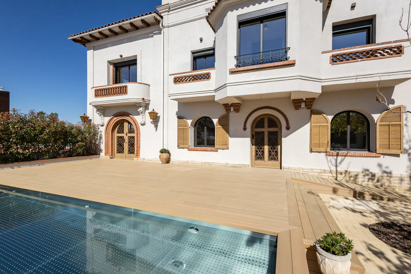 Exterior of a white two-story house with a pool and wooden deck. Arched doors and windows with shutters. Blue sky.