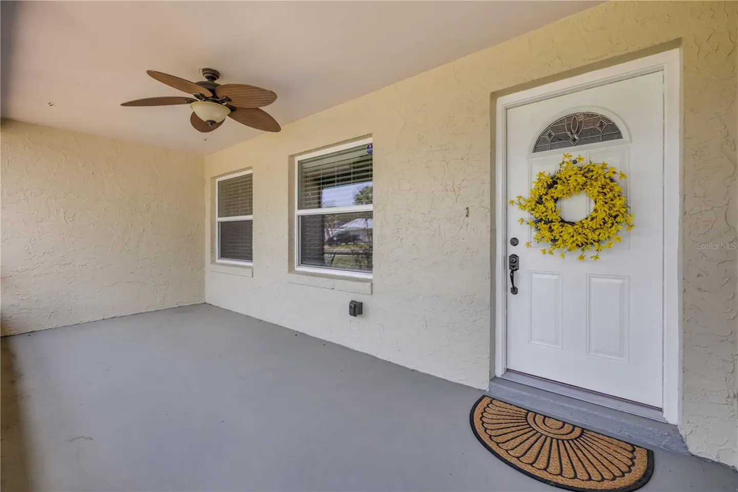 Covered porch with a white front door adorned with a yellow wreath. A ceiling fan, two windows, and a welcome mat complete the scene.