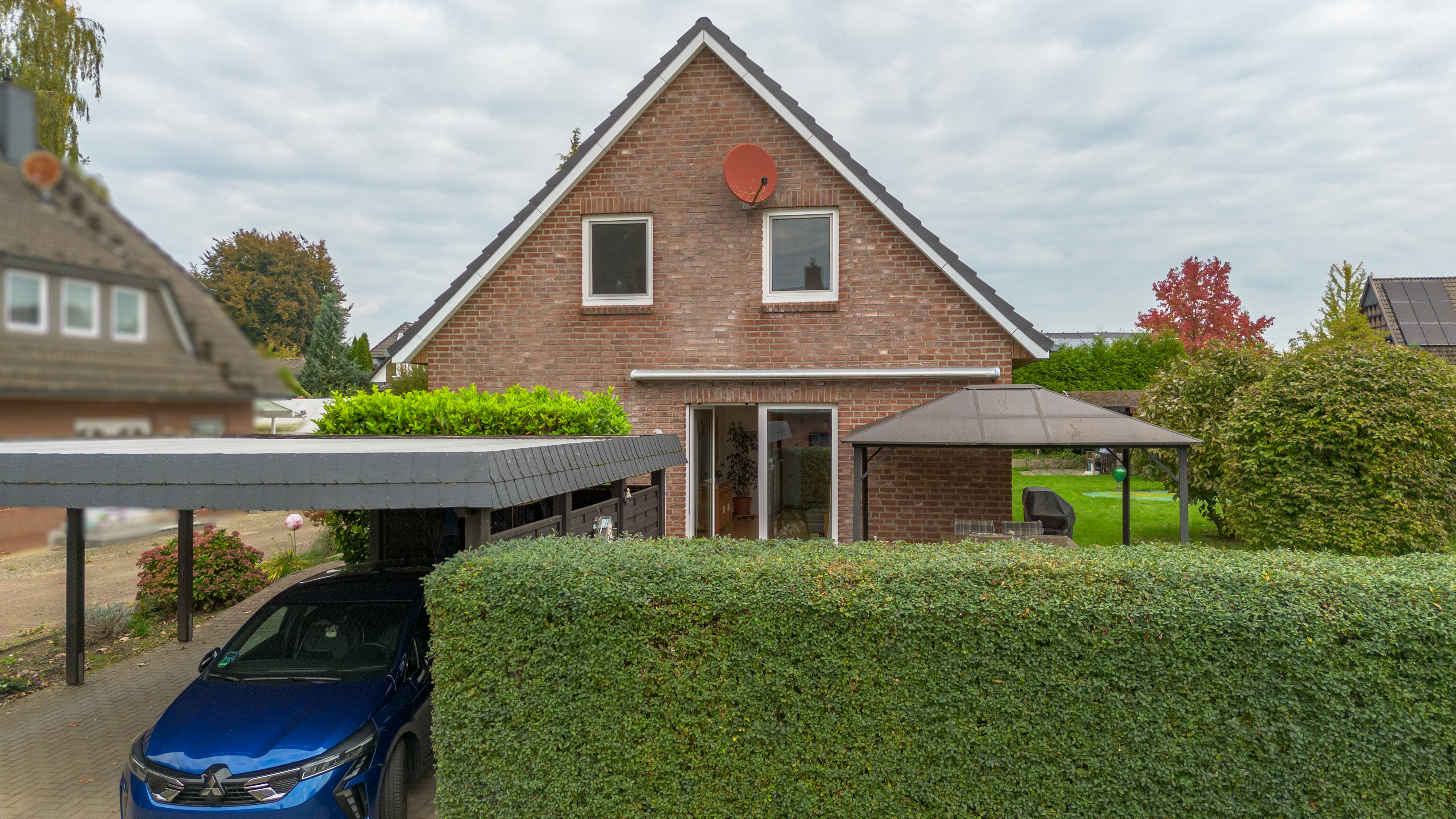 A two-story brick house with a blue car under a carport, a green hedge, and a gazebo in the backyard on a cloudy day. [AI-generated text]