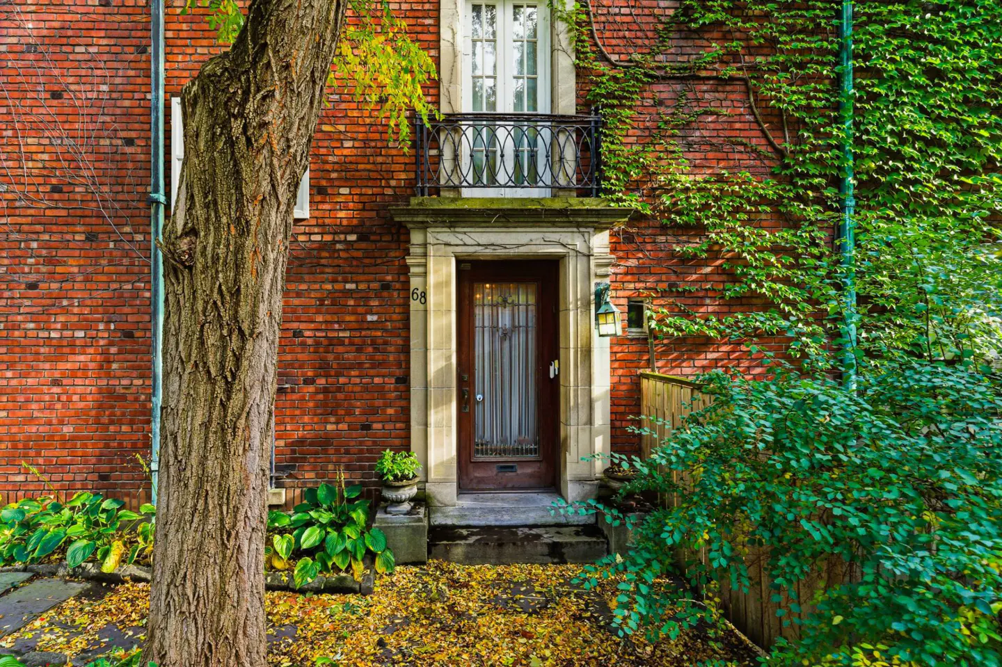 Red brick house with a brown door and a small balcony. Green ivy climbs the walls. A tree stands to the left.