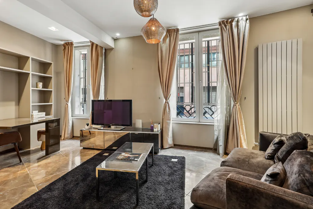 Modern living room with a brown sofa, black rug, glass coffee table, TV console, desk, and beige curtains over windows.