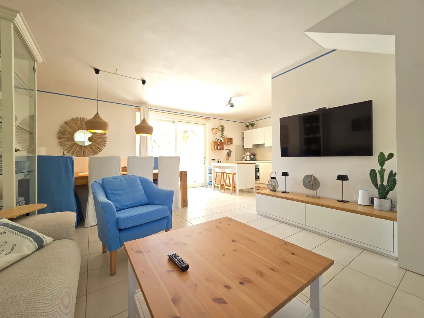 Bright, open living space with a blue armchair, wooden table, and TV. Kitchen visible in the background.