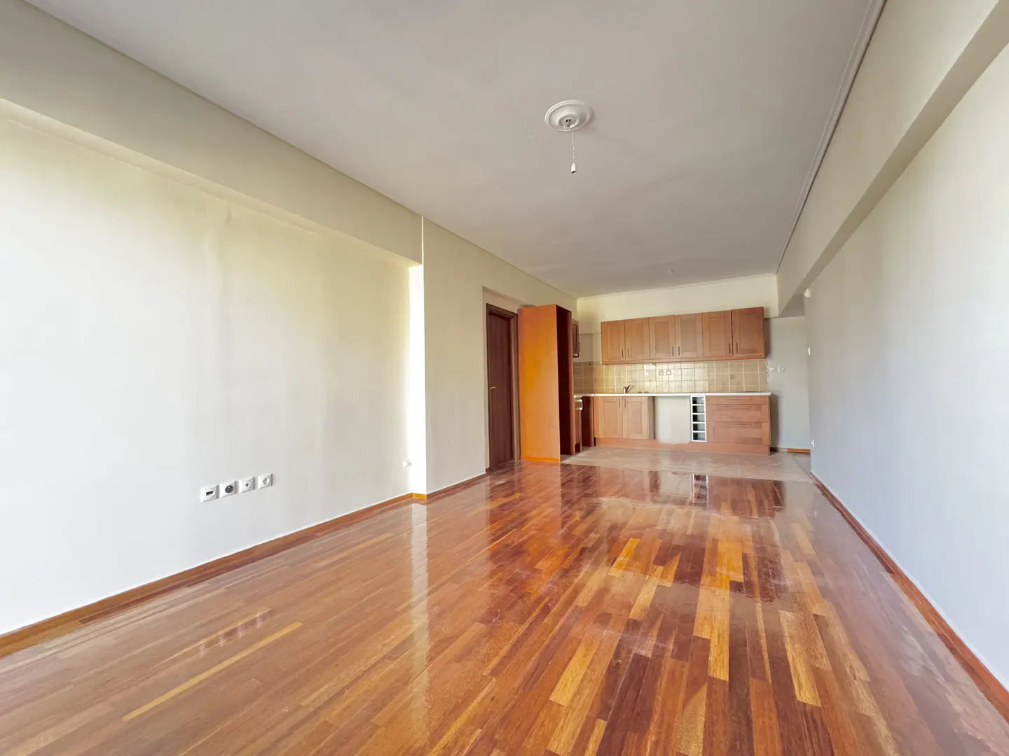 Bright, empty apartment with hardwood floors, white walls, and a kitchen with wood cabinets in the background.