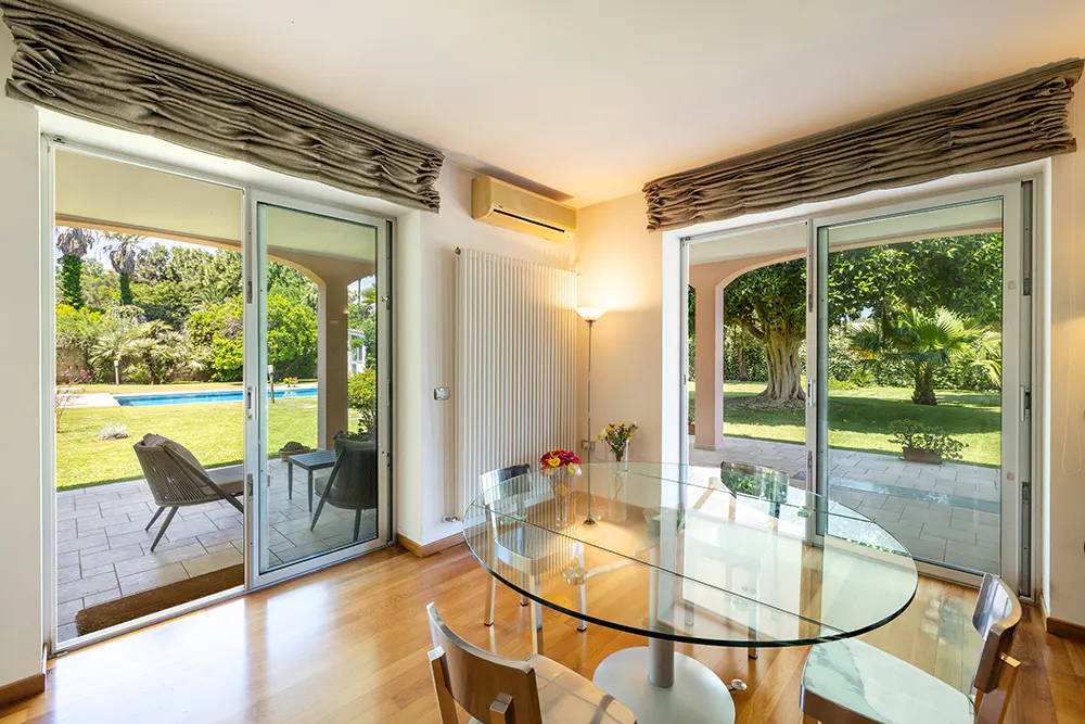 Dining room with glass table, wooden floors, and sliding doors opening to a patio, garden, and swimming pool.
