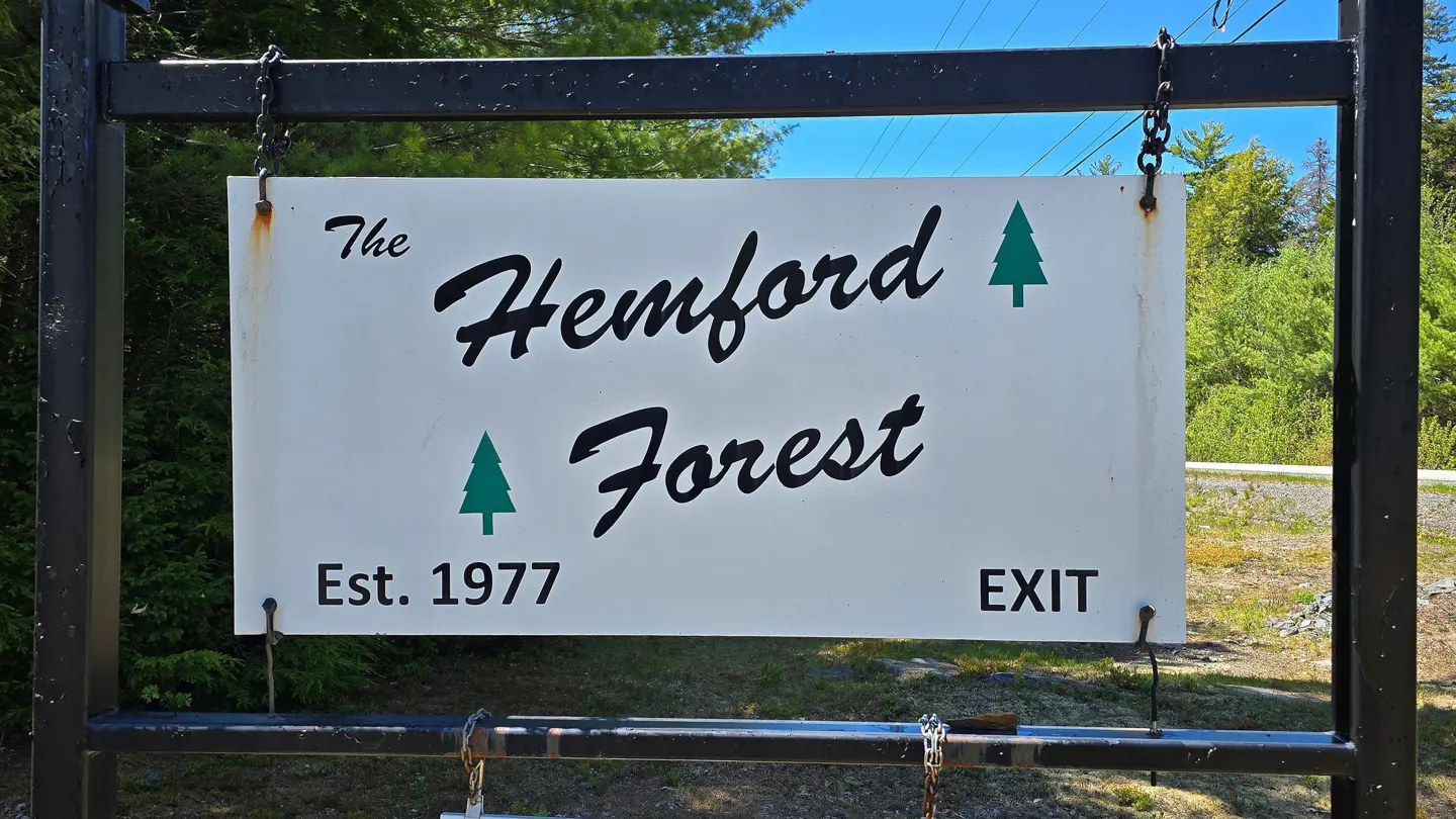 Sign for The Hemford Forest, established in 1977, with green tree icons, hanging from a black metal frame.