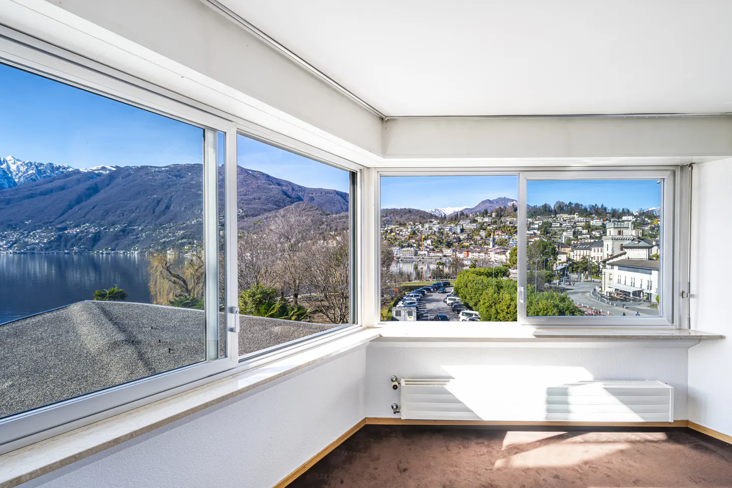 Bright room with large windows showing a lake, mountains, and a town under a blue sky.