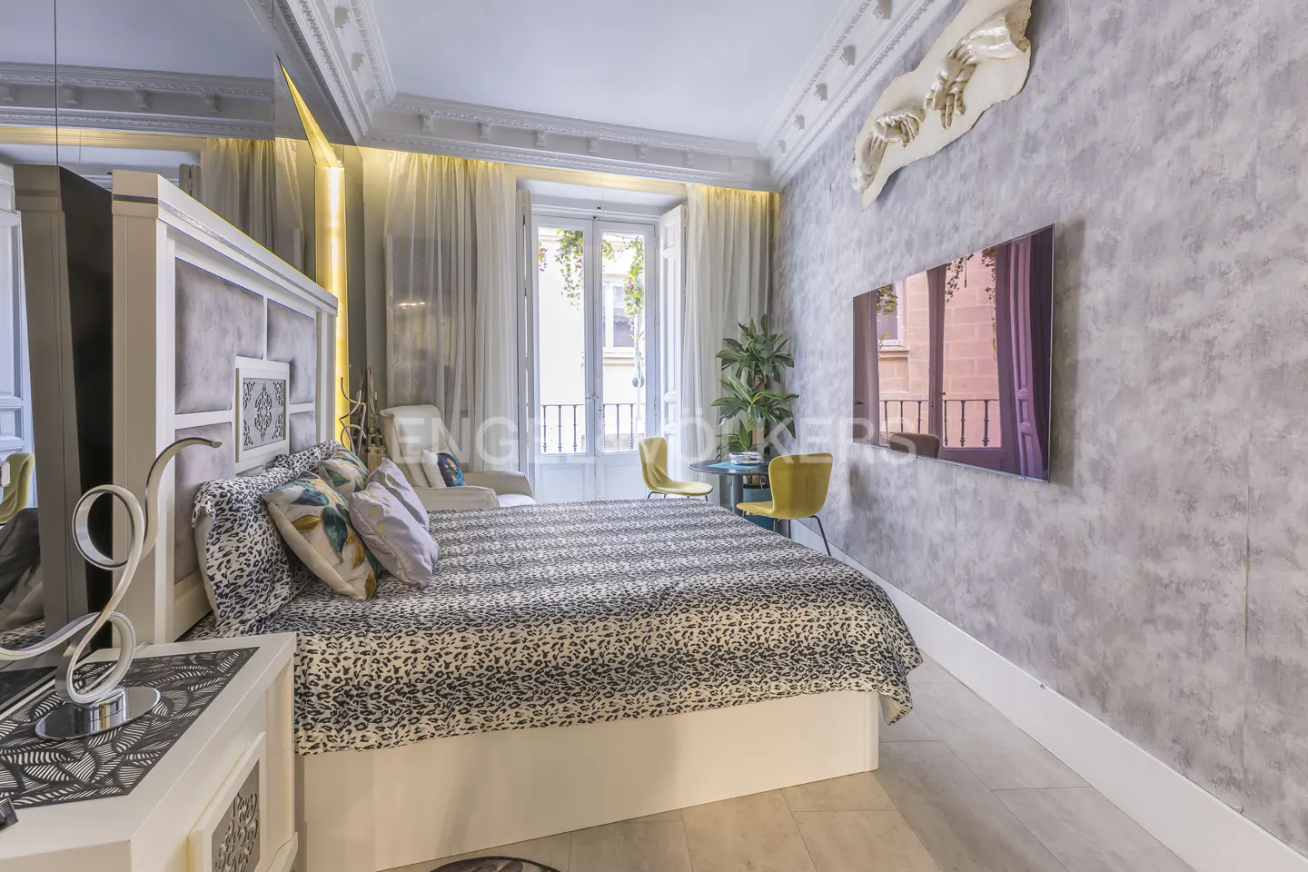 A bedroom with a bed covered in a leopard-print blanket, a TV on the wall, and a balcony with a view of the city.
