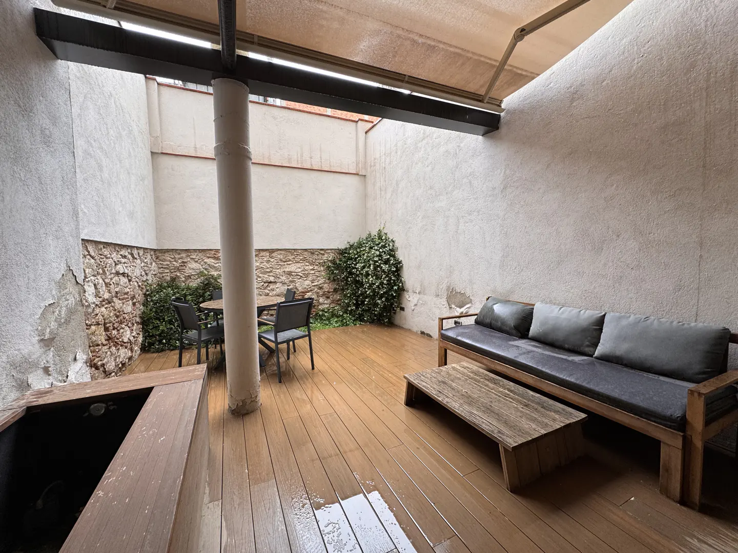 Outdoor patio with wood decking, stone walls, and a neutral color scheme. A sofa, coffee table, and dining set furnish the space.