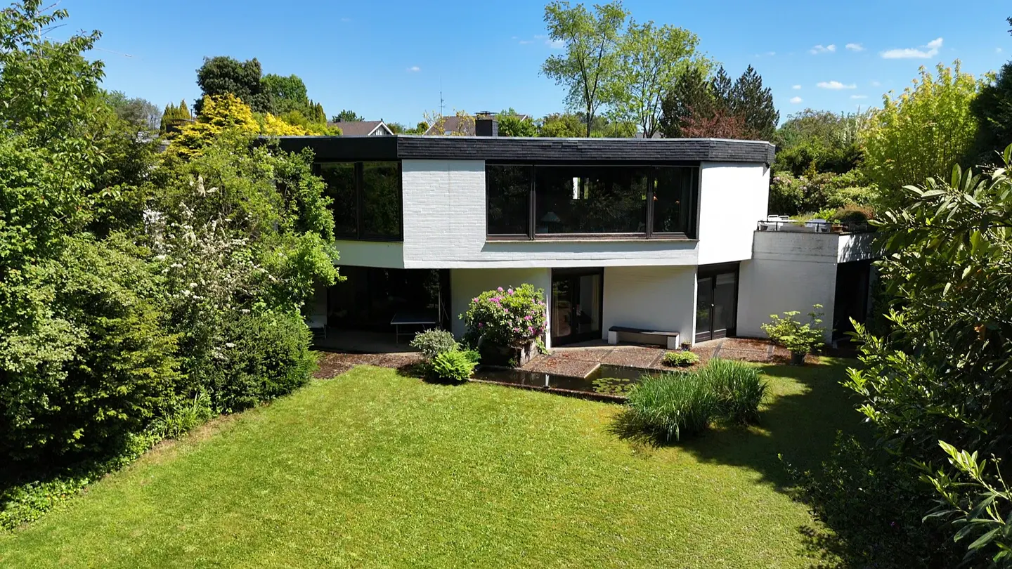 Modern white two-story home with large windows, surrounded by lush green trees and a landscaped garden with a small pond.