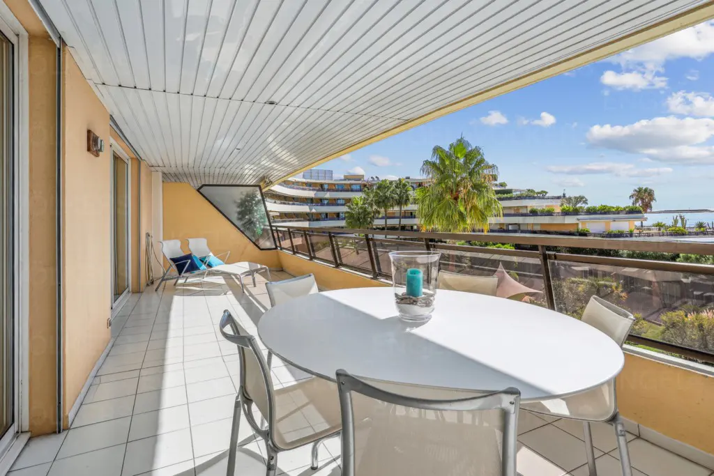 Balcony with white tile floor, table, chairs, and lounge chairs. Palm trees and blue sky are visible in the background.
