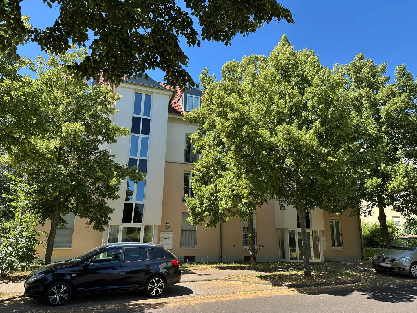 Apartment building with white and beige facade, surrounded by green trees under a blue sky. Two cars are parked in front.