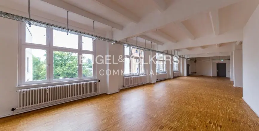 Empty office space with light wood floors, white walls, large windows, radiators, and exposed ceiling cable trays.