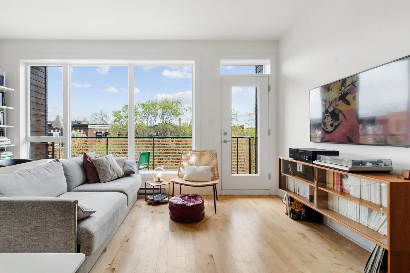 Bright living room with a gray sofa, wood floors, and a large window overlooking a balcony. A TV hangs above a wooden media console.