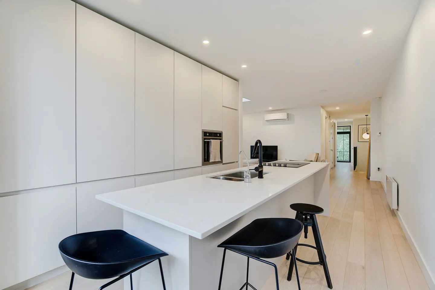 A modern kitchen with white cabinets, a white countertop island, and black bar stools. The floor is light wood.