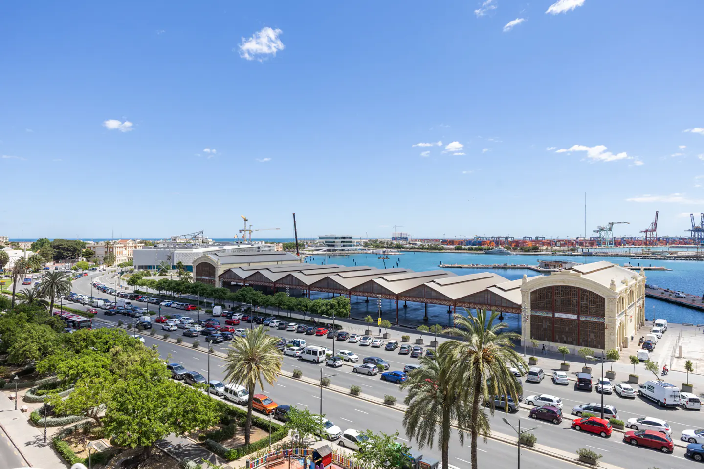 Cityscape view of Valencia, Spain, featuring a busy street, palm trees, and the port in the background under a blue sky.