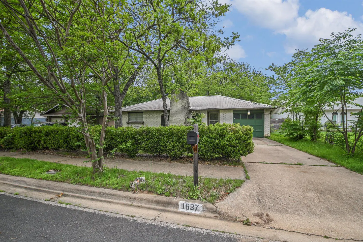 Front view of a one-story white brick house with a green garage door and trimmed hedges. A mailbox stands in front of the house.